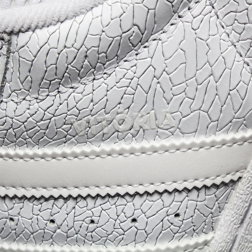 Close-up of a white sneaker with a cracked texture, featuring "VITORIA MENDONCA" embossed on the side. The design includes a flexible cupsole for added comfort and style, from the ADIDAS X VITORIA MENDONCA SUPERSTAR ADV CORE WHITE / WHITE REFLECTIVE collection by ADIDAS.