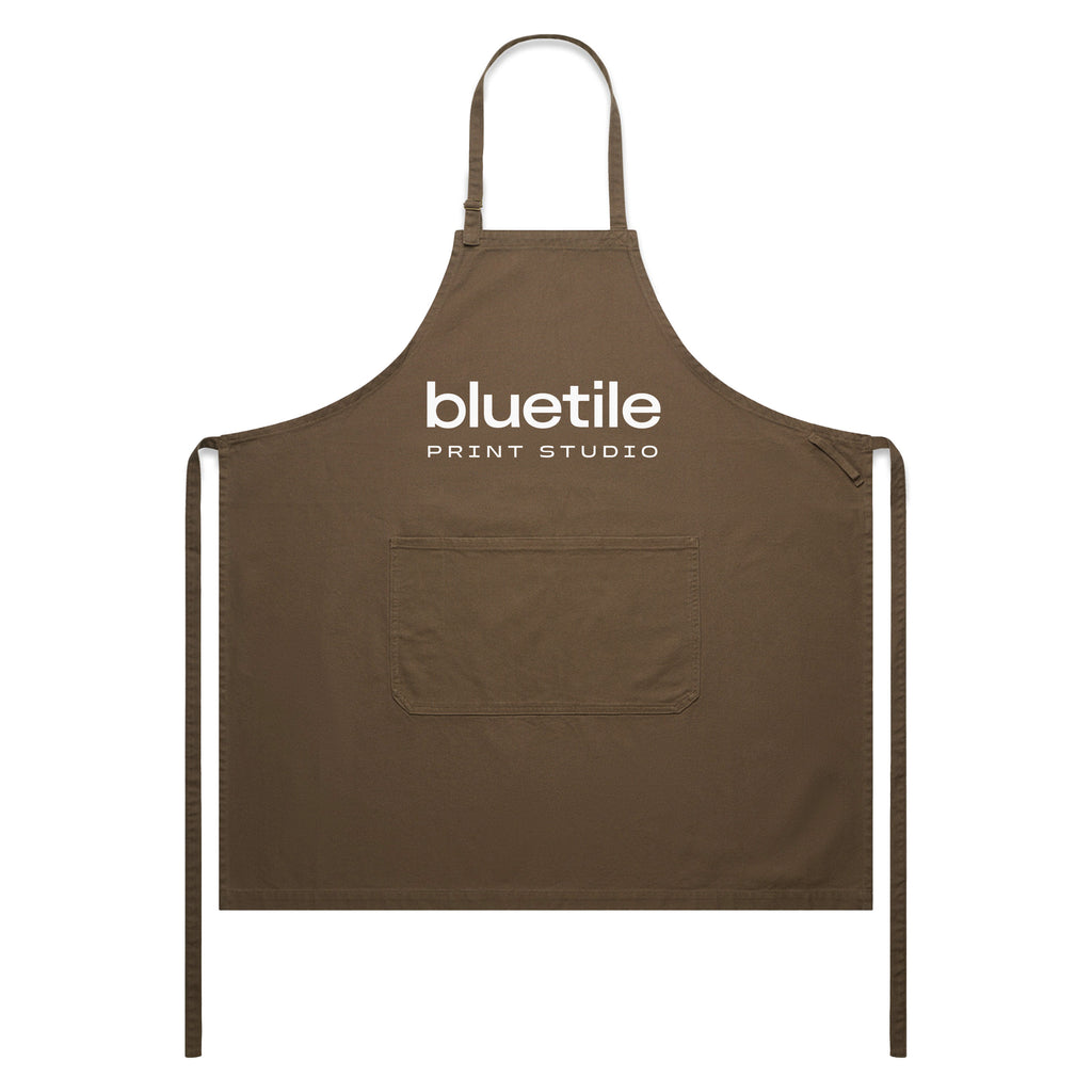 The BLUETILE PRINT STUDIO CANVAS APRON WALNUT by Bluetile Skateboards features a neck loop, adjustable strap, waist ties, and a central pocket for convenience. Crafted from 100% cotton, it offers both comfort and durability, with "bluetile PRINT STUDIO" printed on the front.