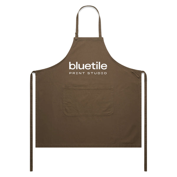 The BLUETILE PRINT STUDIO CANVAS APRON WALNUT by Bluetile Skateboards features a neck loop, adjustable strap, waist ties, and a central pocket for convenience. Crafted from 100% cotton, it offers both comfort and durability, with "bluetile PRINT STUDIO" printed on the front.