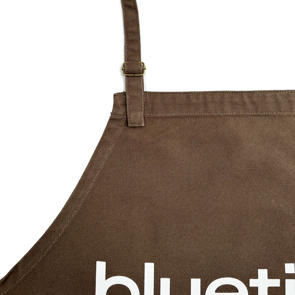 Close-up of a brown canvas apron made from 100% cotton, featuring an adjustable strap. Part of the word "Bluetile" is visible in crisp white text.