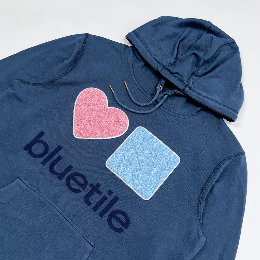 BLUETILE CHENILLE HEART & SQUARE HOODIE SEASIDE by Bluetile Skateboards is a navy blue hoodie made from organic cotton, featuring a pink heart and light blue square design with the word "bluetile" underneath.