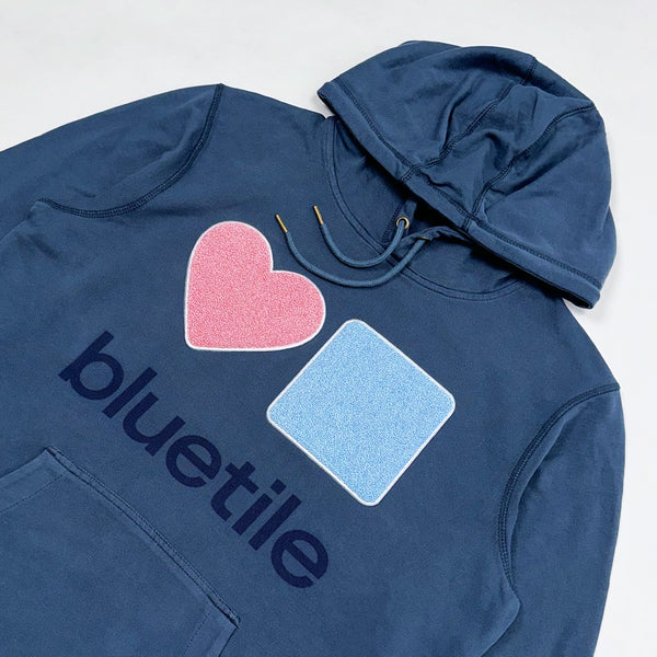 BLUETILE CHENILLE HEART & SQUARE HOODIE SEASIDE by Bluetile Skateboards is a navy blue hoodie made from organic cotton, featuring a pink heart and light blue square design with the word "bluetile" underneath.