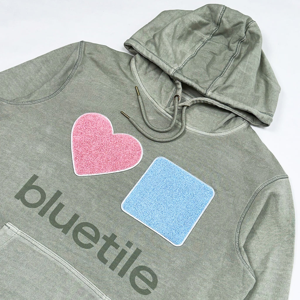 The BLUETILE CHENILLE HEART & SQUARE HOODIE SAGE by Bluetile Skateboards features "bluetile" text, along with a pink heart and a blue square patch on the front. Made from anti-pill fleece, it's ideal for skateboard enthusiasts who appreciate both style and durability.