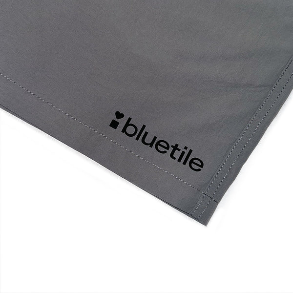 A close-up of the BLUETILE LOGO SWIM SHORTS SHADOW by Bluetile Skateboards shows the gray quick-dry fabric with "bluetile" and a black heart logo printed on the corner.