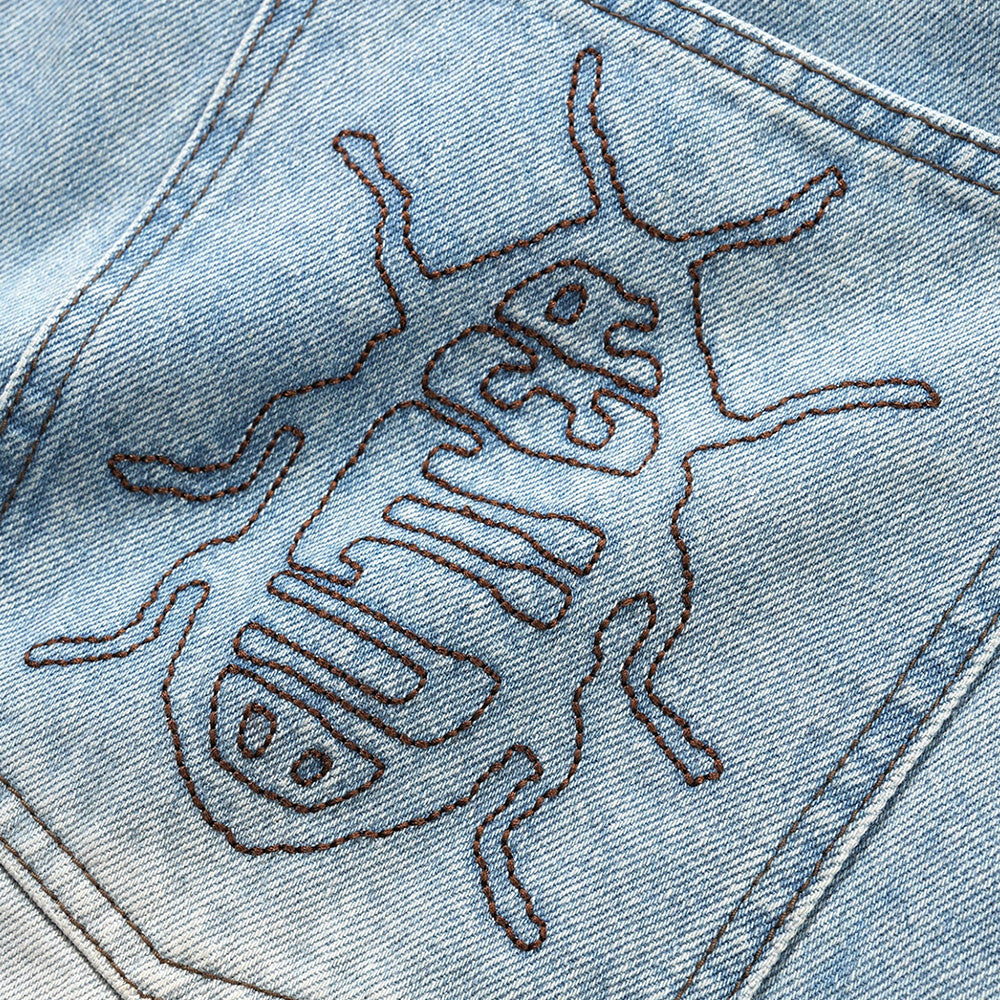 Close-up of the distressed indigo BUTTER ROACH DENIM SHORTS by Carpet Co., showing light blue denim with a stitched geometric cartoon bug design.