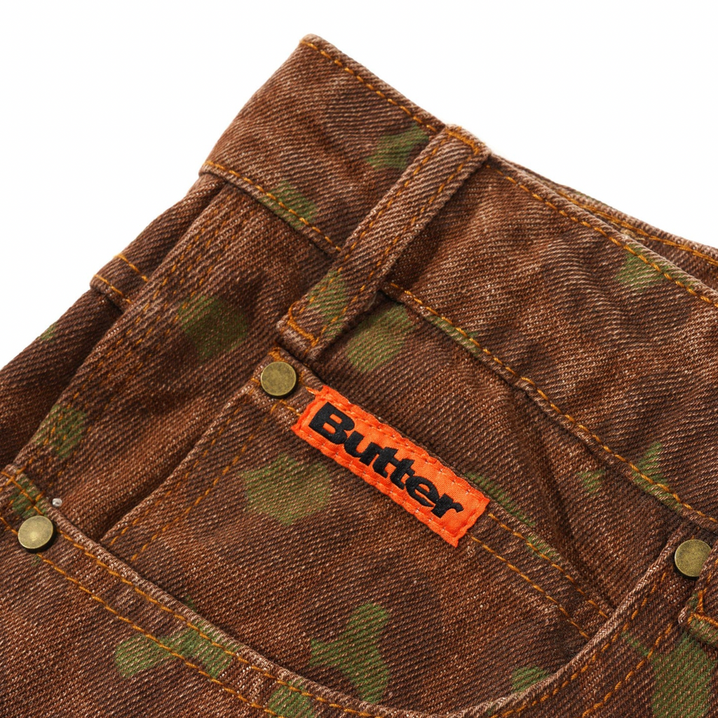 Close-up of the BUTTER GOODS LEOPARD DENIM JEANS RUST with a distinct camouflage pattern and an orange "Butter Goods" tag sewn onto the back pocket, featuring a subtle laser graphic.