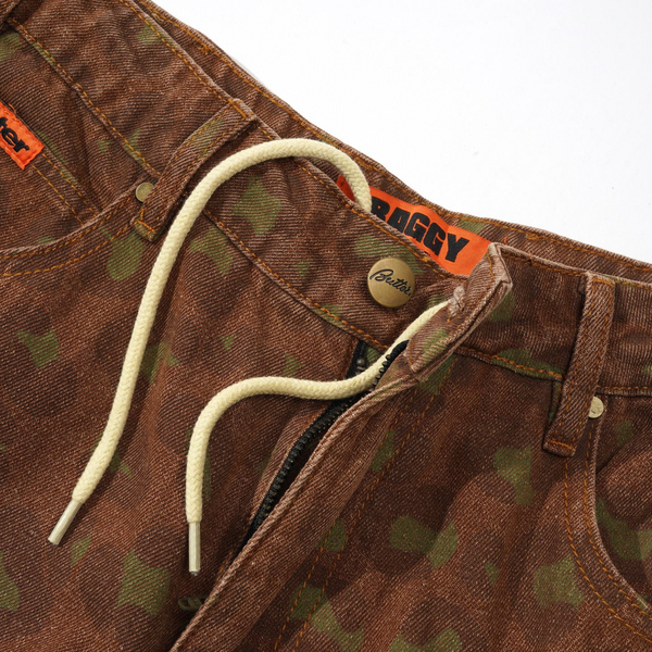 Close-up of a pair of rust-colored leopard print denim jeans with an orange label that reads "BUTTER" and white drawstrings hanging from the waistband. Reminiscent of baggy denim jeans, these pants feature a metal button and a zipper fly.