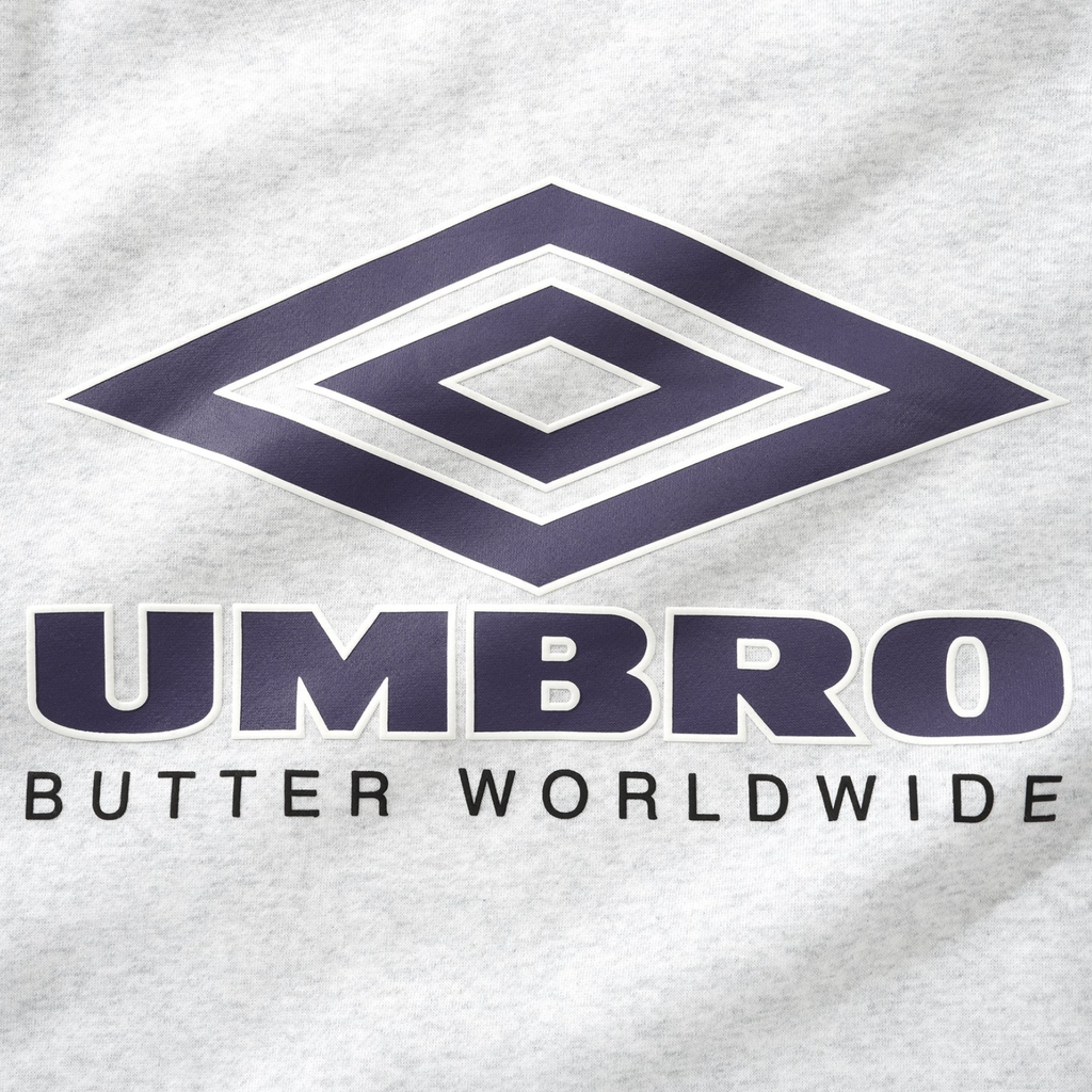 Close-up of a cotton blend fabric featuring the Umbro logo above the text "BUTTER WORLDWIDE" on the Butter Goods x Umbro Diamond Logo Crewneck Ash, highlighting detailed embroidery artwork.