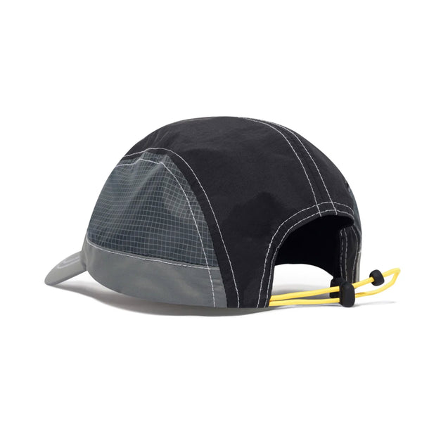 The Butter Goods x Umbro Training Cap Black is a black and gray trucker cap featuring a grid pattern, an adjustable yellow cord, and a curved bill, complemented by mesh back panels.