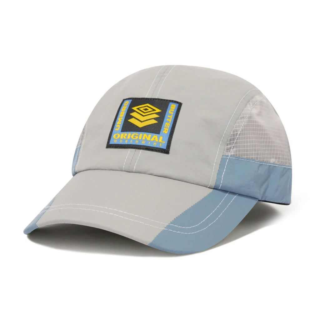 The BUTTER GOODS X UMBRO TRAINING CAP CEMENT by Butter Goods is a gray and blue cap showcasing a black and yellow "Original" patch on the front, seamlessly blending classic style with contemporary flair.