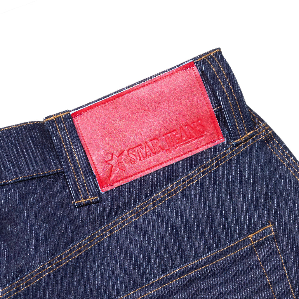 Close-up of 100% cotton blue denim jeans by Carpet Co., featuring orange stitching and a red leather "CARPET CO. C-STAR JEANS RAW DENIM" patch on the waistband.