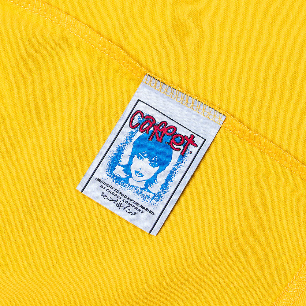 A white tag with blue and red graphics, featuring an illustrated face and "CORRECT," is sewn onto yellow cotton fabric from the Carpet Co. CAT TEE YELLOW, emphasizing its screen printed details.