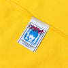 A white tag with blue and red graphics, featuring an illustrated face and "CORRECT," is sewn onto yellow cotton fabric from the Carpet Co. CAT TEE YELLOW, emphasizing its screen printed details.