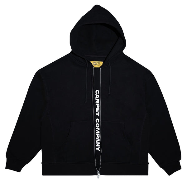 The Carpet Co. FAKE ZIP UP HOODIE BLACK features a double zip design, hood, front pockets, and "CARPET COMPANY" printed in white along the zipper—a stylish and comfortable choice for daily wear.