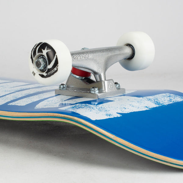 A skateboard with white wheels and silver trucks on a FOUNDATION THRASHER COMPLETE BLUE deck with white splatter design.