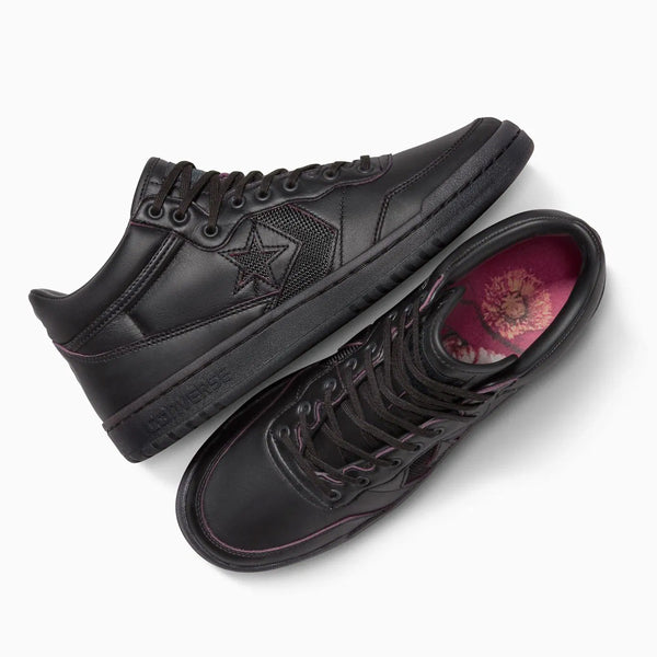 The CONVERSE CONS Remy Taveira Fastbreak Pro Mid features a nubuck upper with a star logo. It comes in all black with floral-patterned insoles, black laces, and one shoe displayed sideways while the other highlights its sole.