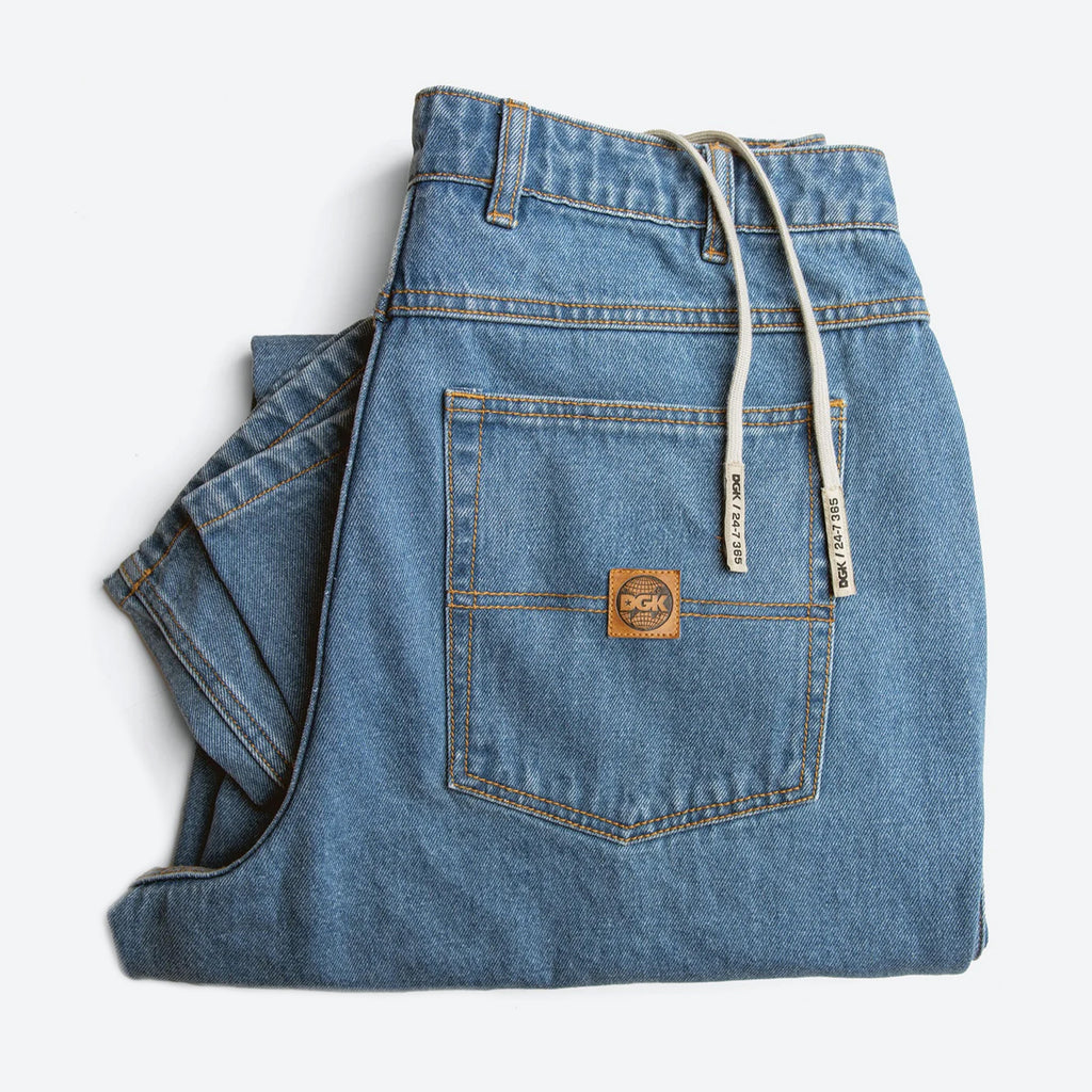 The DGK MUNI DENIM JEAN WASHED INDIGO offers a comfortable baggy fit in a folded blue denim design. It is equipped with a YKK zipper fly, beige drawstrings, and features a brown leather patch labeled "DGK" on the back pocket.