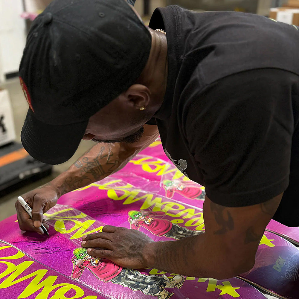A person in a black cap and shirt uses a silver marker to sign the colorful DGK Stevie Williams Cheesin Autographed poster by DGK, which features cartoon graphics like those on an 8.38-width skateboard.