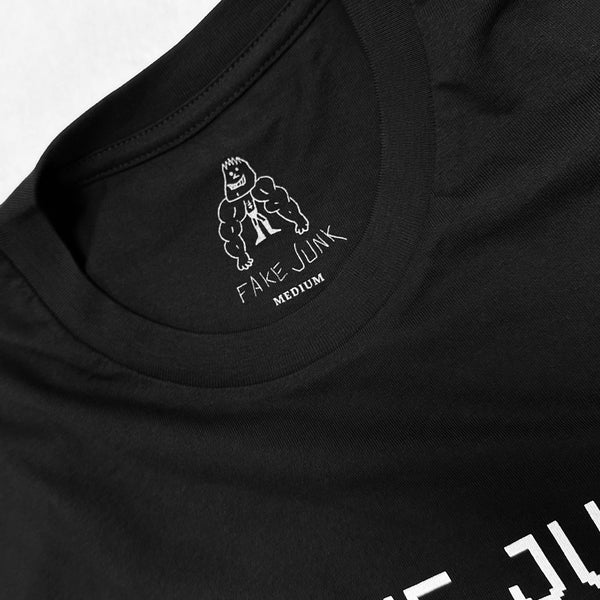 Close-up of a black t-shirt's inside label, crafted from combed cotton, featuring a cartoon muscular figure with "FAKE JUNK PROTECT TEE BLACK" written below and the size "Medium" indicated.