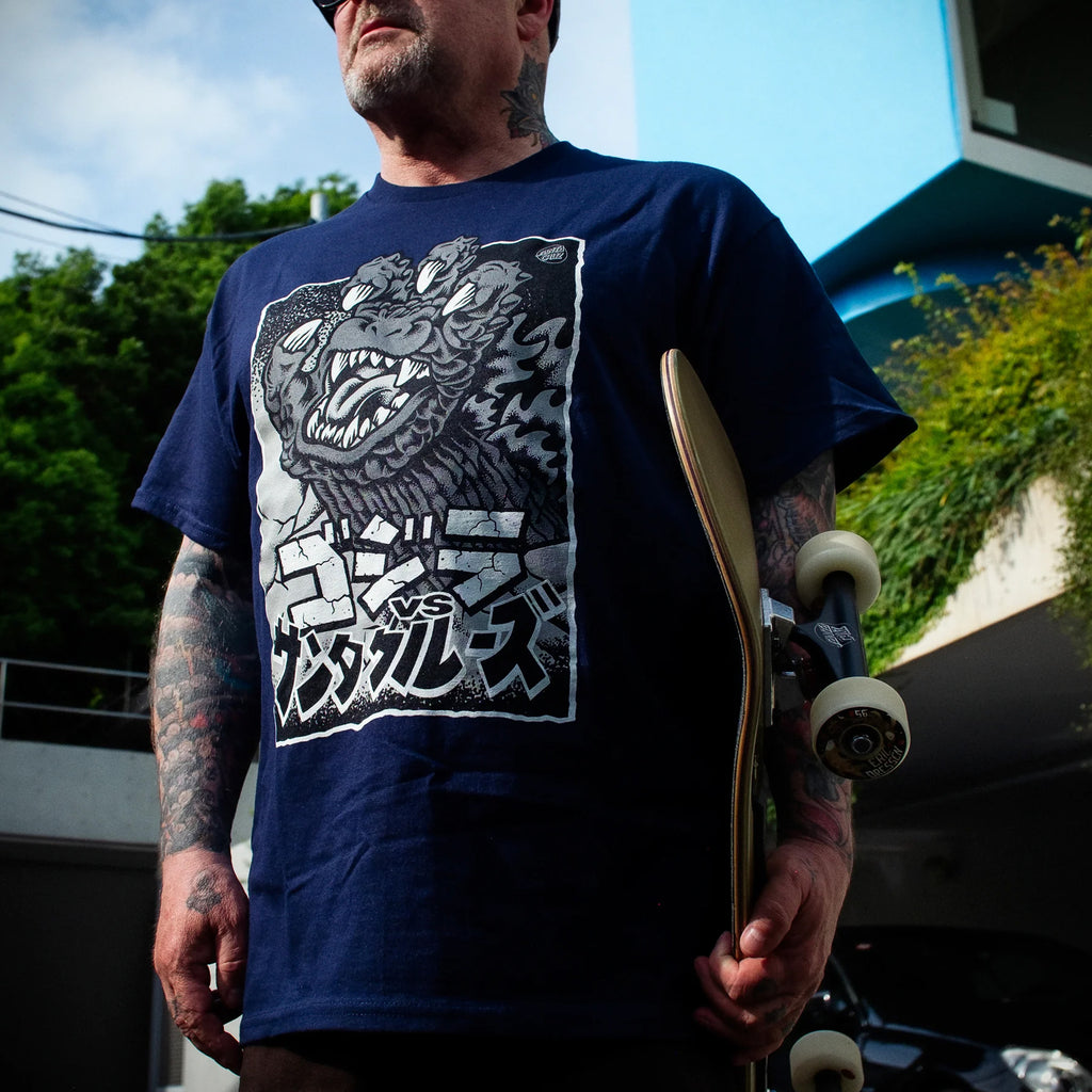 A person wearing a SANTA CRUZ X GODZILLA HAND TEE NAVY holds a skateboard outdoors.