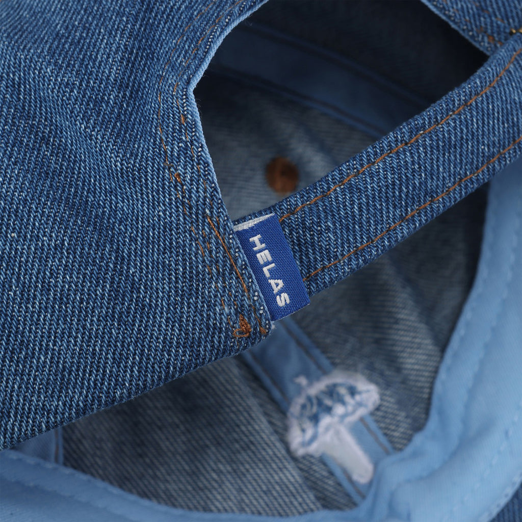 Close-up of a light blue denim garment showing a tag with the text "HELAS CLASSIC DENIM CAP LIGHT BLUE" and an embroidered umbrella design.