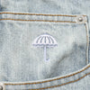 Close-up of a denim fabric with a small embroidered white umbrella above a pocket, featuring visible stitching and metal rivets. The detail complements the casual aesthetic of the HELAS DIEU GREC JEAN SHORT LIGHT BLUE from HELAS, ideal for everyday wear.