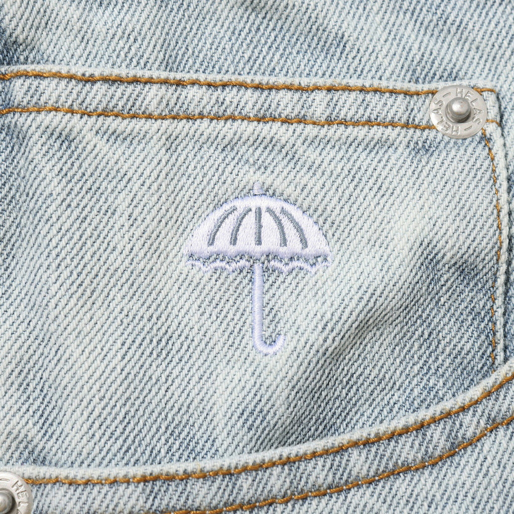 Close-up of a denim fabric with a small embroidered white umbrella above a pocket, featuring visible stitching and metal rivets. The detail complements the casual aesthetic of the HELAS DIEU GREC JEAN SHORT LIGHT BLUE from HELAS, ideal for everyday wear.