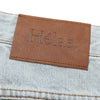 Close-up of a brown leather patch with the word "Hélas" embossed on it, attached to light blue denim fabric, featuring an embroidered logo. Product: HELAS DIEU GREC JEAN SHORT LIGHT BLUE by HELAS.