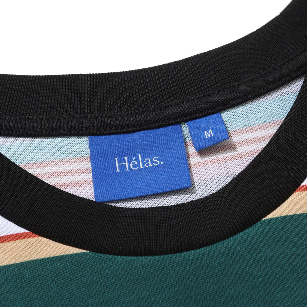 Close-up of a striped shirt collar with a black edge. A blue tag with the brand name "Hélas" and a size tag marked "M" are visible, showcasing the HELAS RAYURES TEE GREEN / BLACK in 100% COTTON. An embroidered umbrella logo adds a stylish touch.