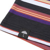 Close-up of a Helas Rayures Tee Purple/Black fabric with horizontal stripes in black, purple, red, yellow, and white, featuring a small tag with an embroidered umbrella logo.