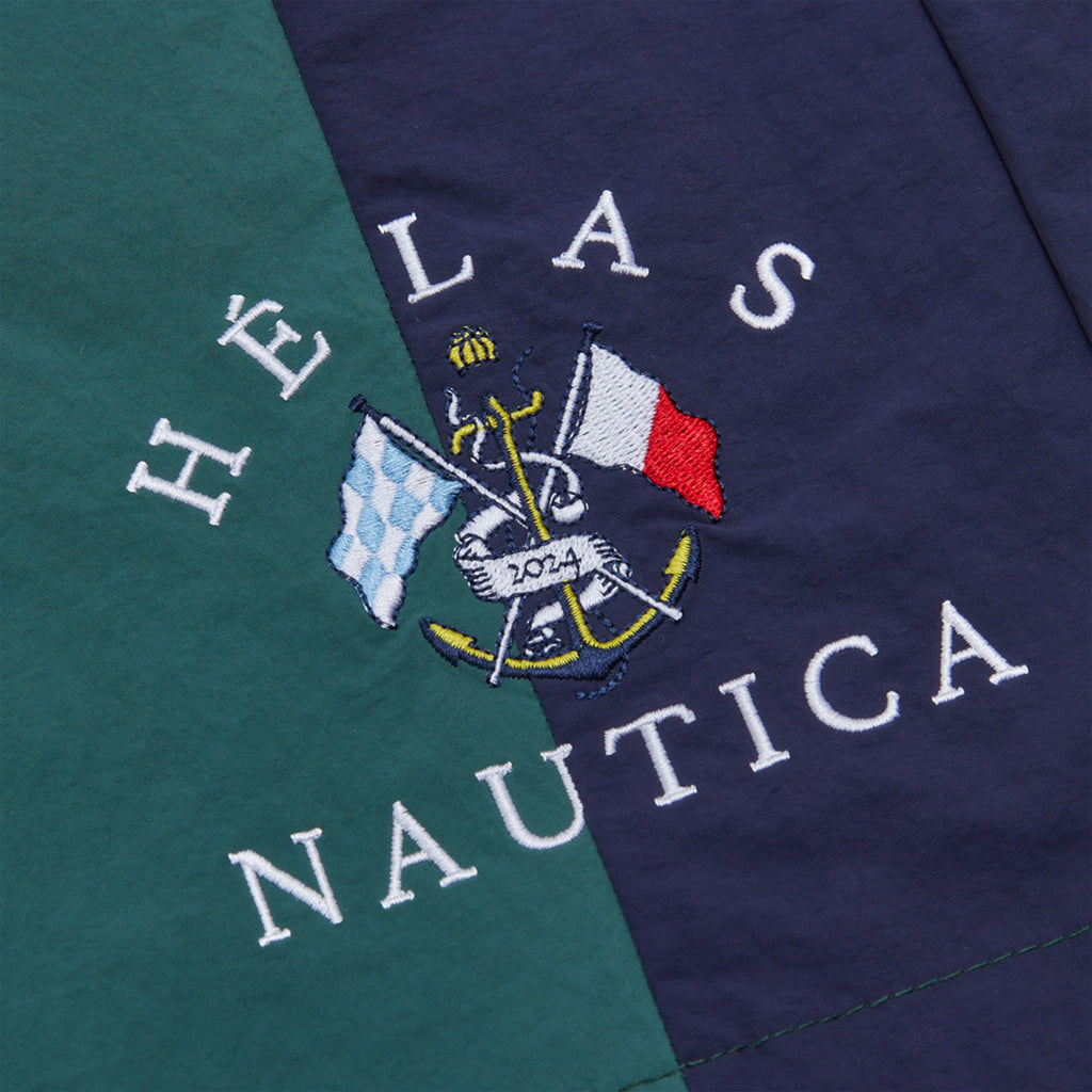 Close-up of the HELAS X Nautica Yannick 8.5" Swim Short in Moss Green showcasing an embroidered logo with the text "HÉLAS NAUTICA," adorned with an anchor and two flags—one checkered and one resembling the French flag—all stitched meticulously, perfect for highlighting a unique detail on these swim shorts.