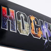 A close-up view of stylized text spelling "HOCKEY" on a skateboard from HOCKEY. Each letter features different graphics including lightning, a doppleganger statue, flames, and smoke. The vibrant colors are brought to life with premium digital print and silver foil highlights.