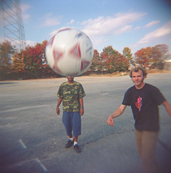 Two men playing with a LIMOSINE MAX PALMER MUNDO soccer ball, with the ball captured in motion close to the camera, in a parking lot with trees in the background.