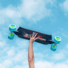 A person extends an arm upwards, holding a LOADED FATHOM 33" COMPLETE longboard against a blue sky with scattered clouds. The skateboard has blue and green wheels.