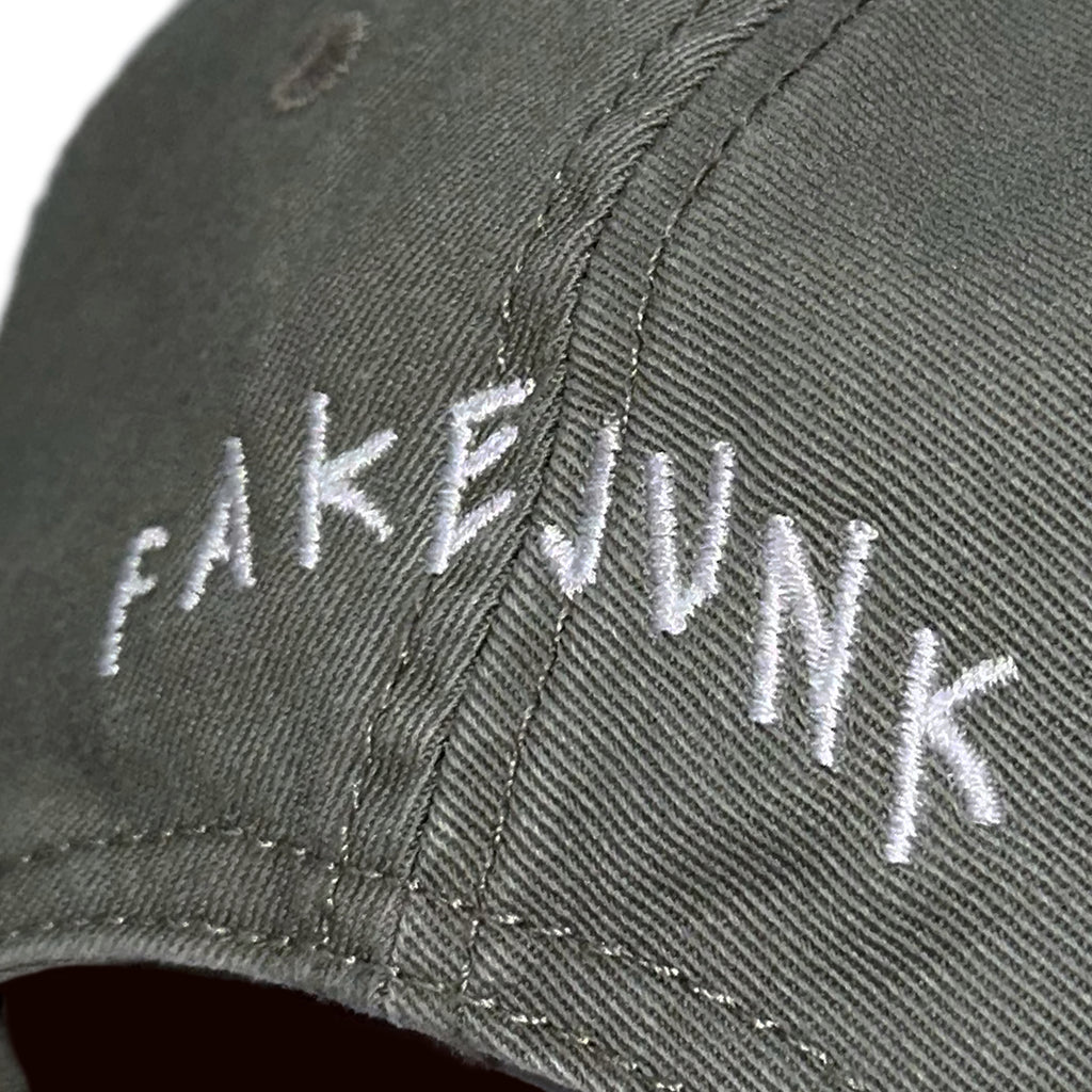 Close-up of a dark green fabric cap, the FAKE JUNK LOGO DAD HAT EUCALYPTUS, with "FAKEJUNK" embroidered in white thread on the front; features an adjustable fastener and is made from 100% cotton.