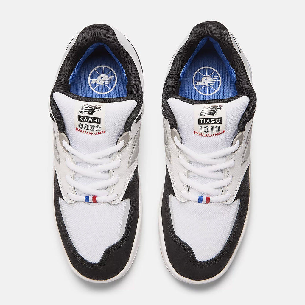 Top-down view of a pair of NB Numeric 1010 Tiago x Kawhi White/Black sneakers, featuring "KAWHI" and "TIAGO LEMOS" labels on the tongues. Inside the shoes, a blue basketball logo is visible, complemented by FuelCell foam for enhanced comfort. Red, white, and blue accents adorn the lace area.