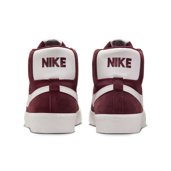 Back view of a pair of NIKE SB BLAZER MID BURGUNDY CRUSH / SUMMIT WHITE sneakers. The word "NIKE" is prominently displayed in maroon on the heel of each shoe.