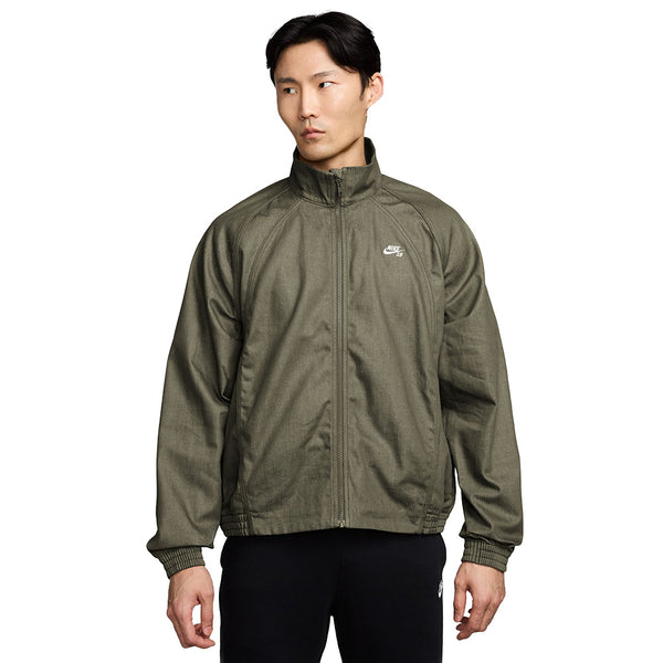 A person wearing the NIKE SB DENIM FULL-ZIP TRACK JACKET in medium olive/white with a small white Nike logo on the chest, standing against a plain white background.