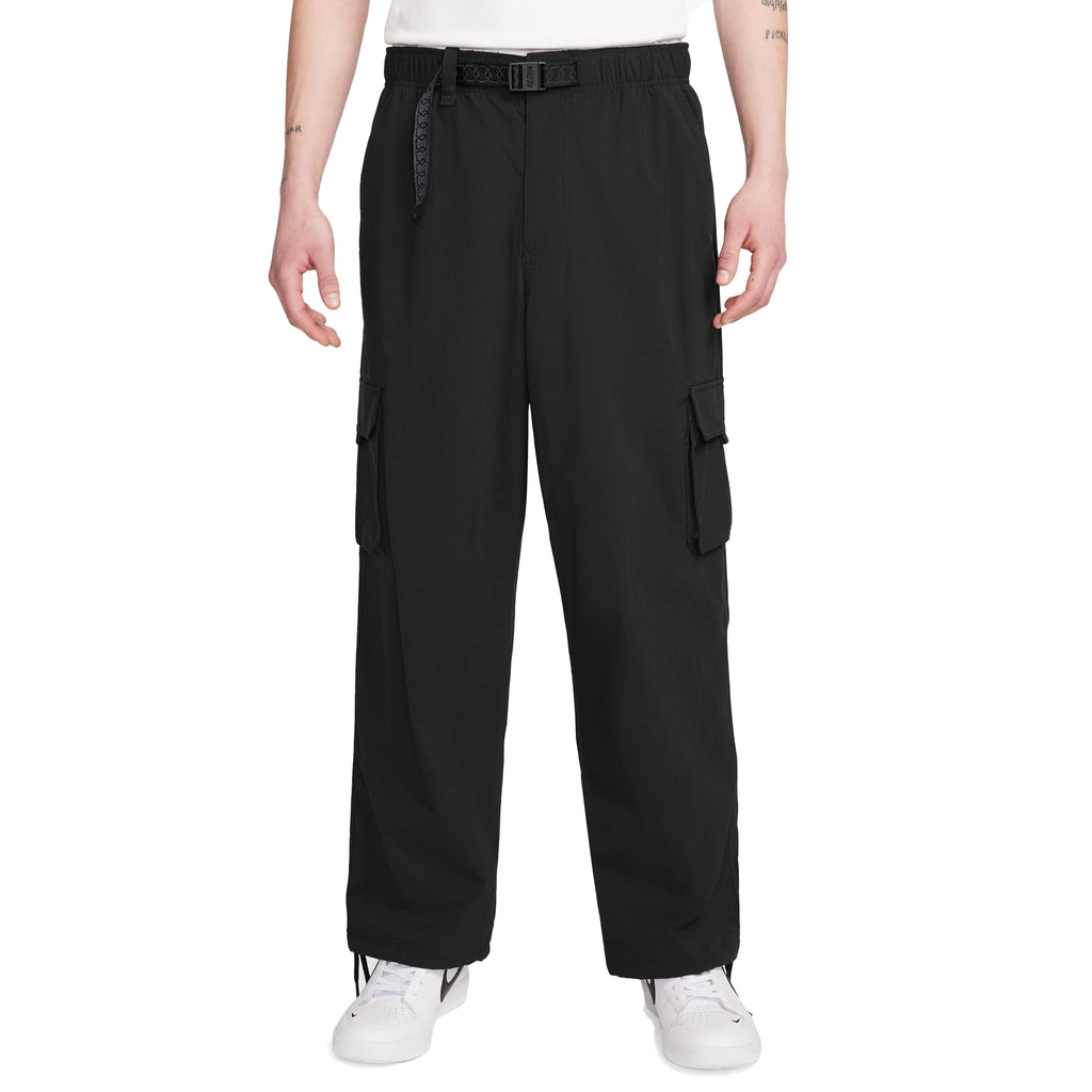 Person wearing Nike SB Kearny Cargo Skate Pants in black with a belt, paired with a white shirt and white Nike SB Kearny sneakers. Standing against a plain white background.