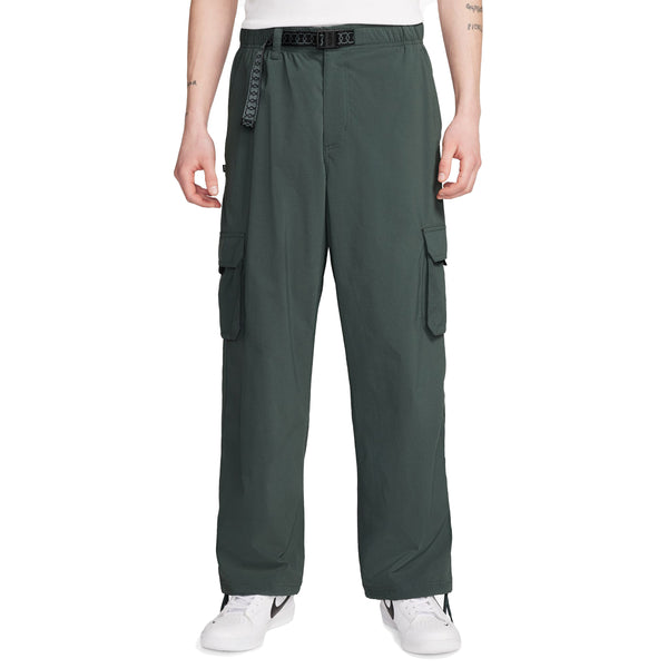 A person wears NIKE SB KEARNY CARGO SKATE PANTS in Vintage Green with a loose fit, paired with a black belt and white sneakers, standing against a white background.