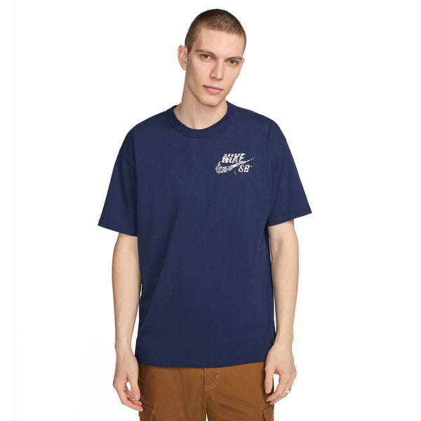 A young man wearing a nike SB YUTO TEE MIDNIGHT NAVY t-shirt with a small white logo on the left chest, paired with brown trousers, stands against a plain background.