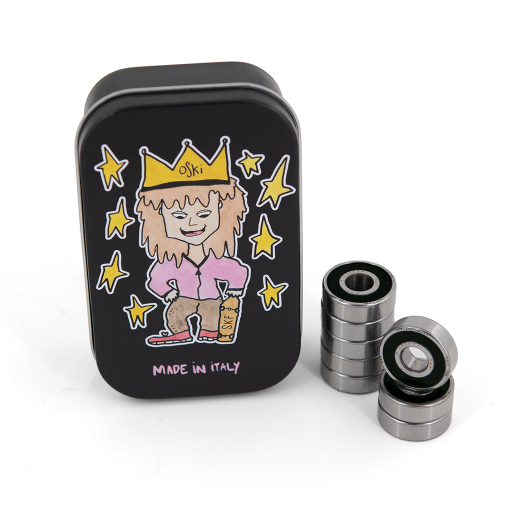 A black rectangular tin featuring a cartoon character and "Made in Italy" text, lies beside the SKF OSKI SIGNATURE BEARING KIT containing five high-quality metal bearings stacked in two columns.