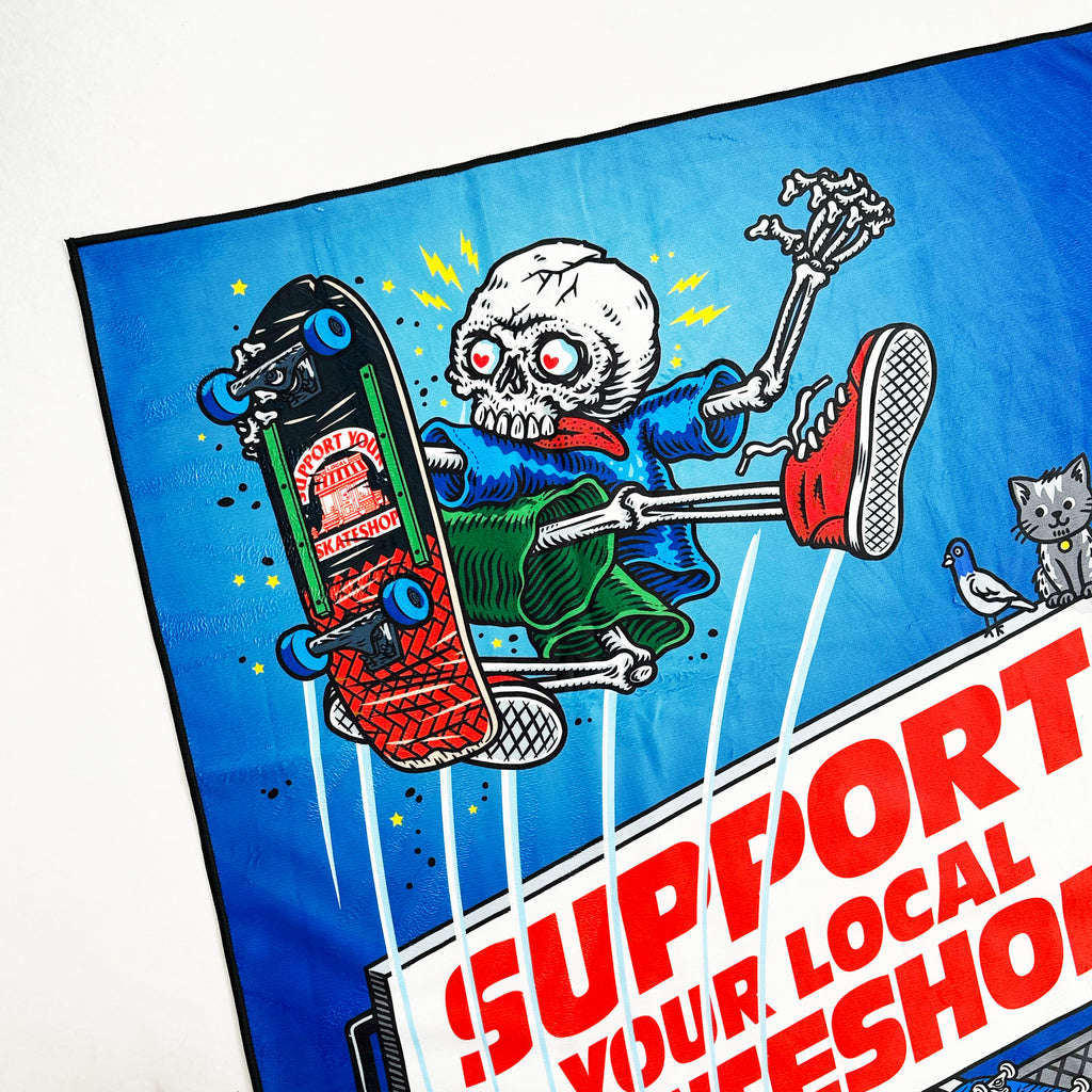 Illustration of a skeleton skateboarding over a billboard with "Support Your Local Skateshop" amid vibrant colors celebrates SKATE SHOP DAY 2025 as a cat and bird join in. This design is featured on the SKATE SHOP DAY 2025 TOWEL by Bluetile Skateboards.