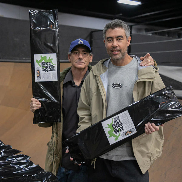 Two men stand side by side at a skate park, each holding a wrapped skateboard deck labeled "GIRL X Skate Shop Day Koston Reissue Blind Bag." The vibrant artwork on one deck channels Mark Gonzales' distinctive style, embodying adventure and creativity in every curve and line.