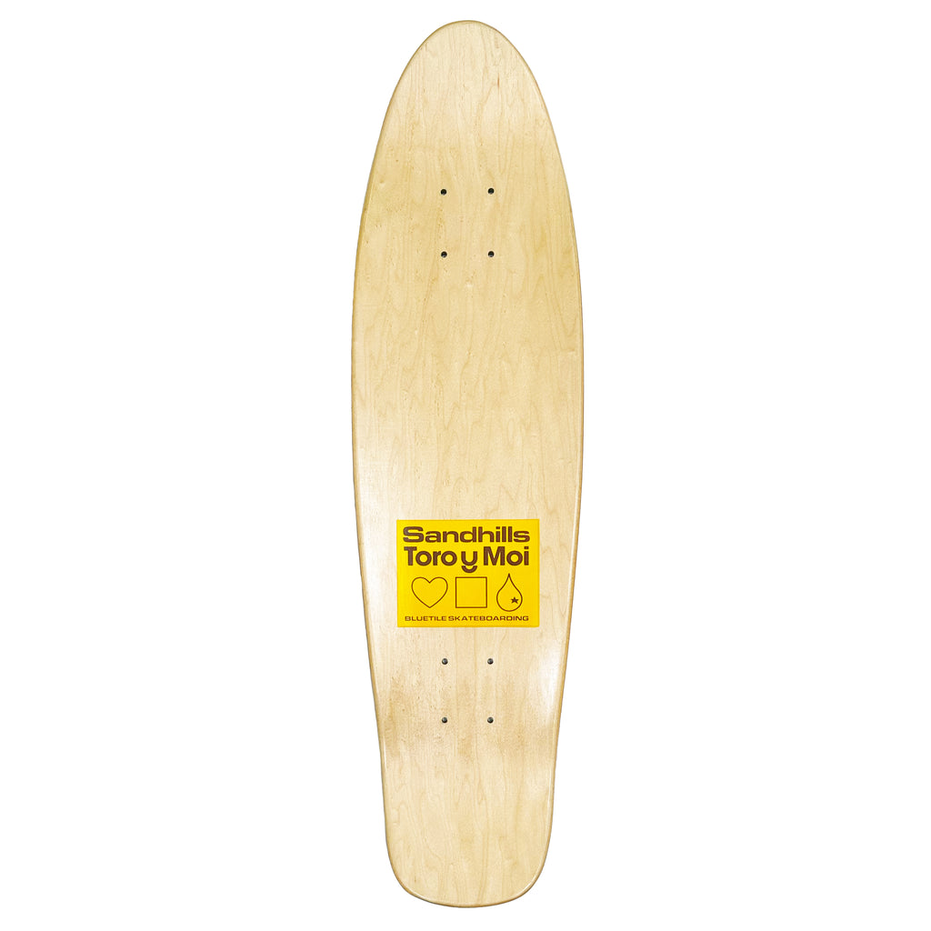 A wooden skateboard with a yellow sticker bearing the Bluetile Skateboards "TORO Y MOI X BLUETILE "SANDHILLS" CRUISER DECK" logo.