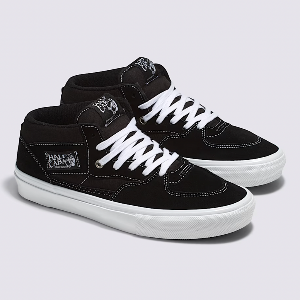 A pair of VANS SKATE HALF CAB BLACK / WHITE sneakers from VANS, featuring white laces, stitching, and soles, are positioned side by side on a light gray background.