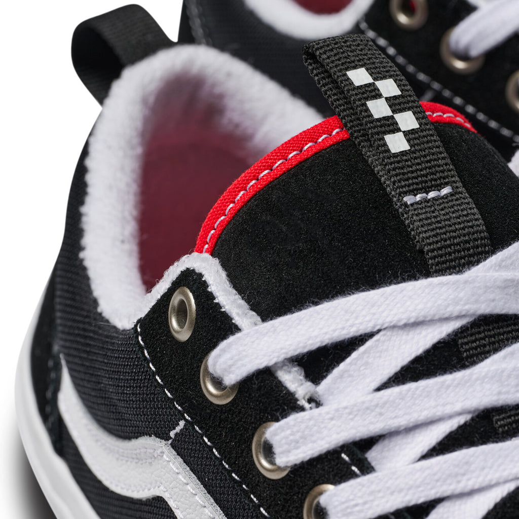 A close-up of the VANS SKATE OLD SKOOL 36+ sneakers in black/white showcases their suede uppers, white soles and laces, red inner lining, and white checks on the tongue tab, capturing the essence of 90s style.