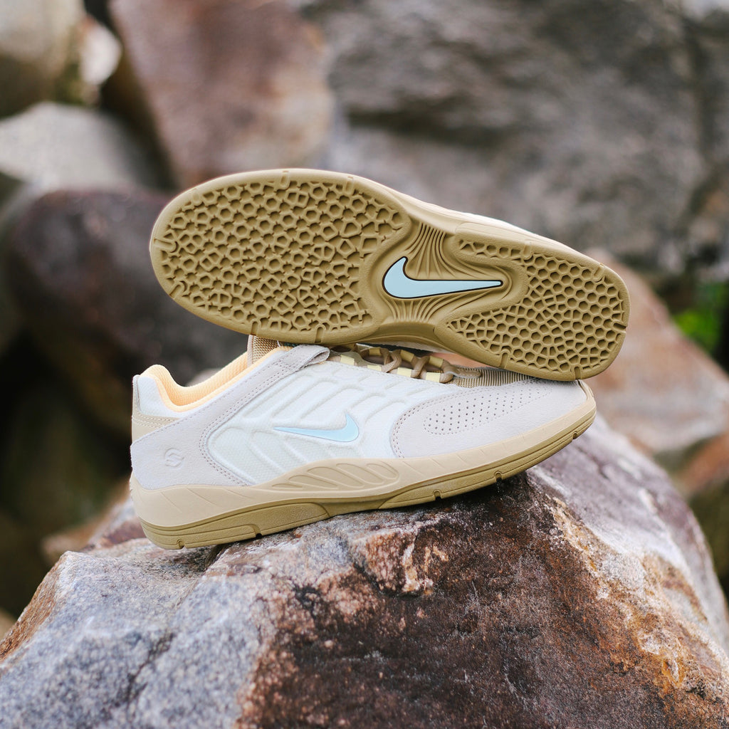 A pair of beige and white NIKE SB VERTEBRAE COCONUT MILK / JADE ICE-SESAME sneakers with light blue accents displayed on a rock, showcasing the textured sole and side design details reminiscent of retro styles by nike.