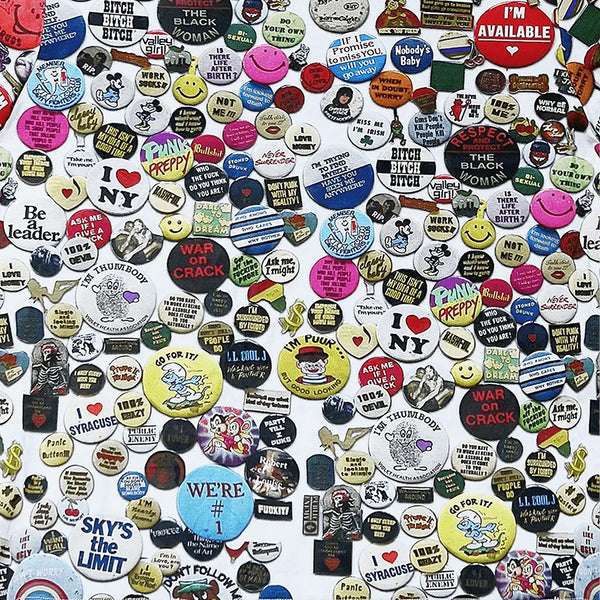 The violet VIOLET PINS TEE is made from 100% cotton and features a crisp white background covered with a vibrant array of vintage-inspired pins, buttons, slogans, logos, smiley faces, and pop culture motifs.
