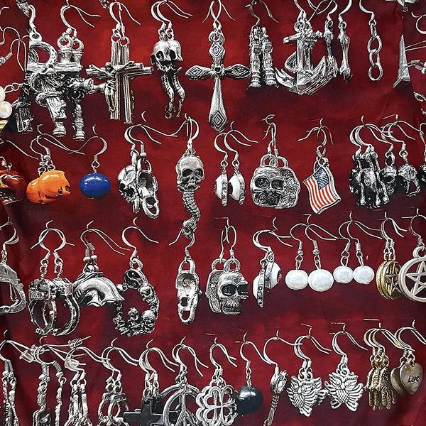 Assorted metal earrings—skulls, crosses, anchors, animals, and symbols—are laid on a red 100% cotton fabric background, evoking the graphic design of the violet VIOLET ST.MARK'S EARINGS TEE.